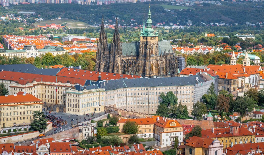 Aerial view of Prague Castle and St. Vitus Cathedral rising above the red-roofed historic buildings of Prague, with green hills and modern cityscape in the background.