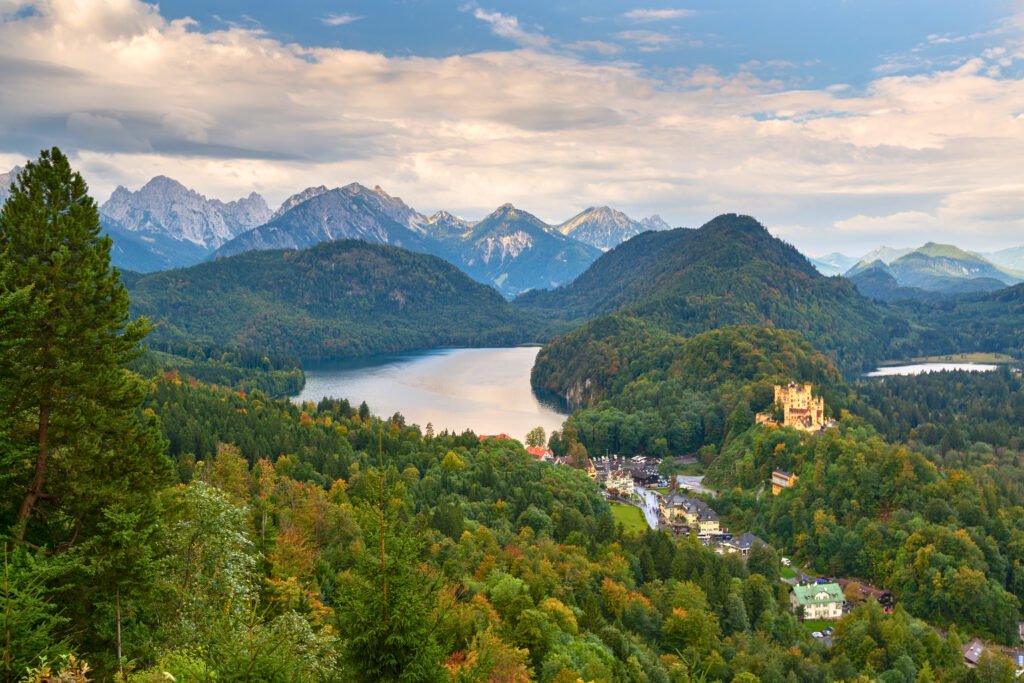 Vue panoramique sur les Alpes bavaroises et le village de Hohenschwangau, en Allemagne, avec le château de Hohenschwangau illuminé au premier plan et un lac pittoresque entouré de forêts verdoyantes et de montagnes en toile de fond.