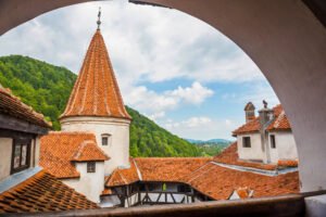 Historic Bran Castle’s red rooftops and turret set amid lush green hills and blue sky.