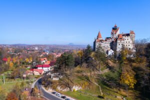 Sunny landscape featuring Bran Castle, legendary Transylvania landmark, rising above colorful autumn village and hills.
