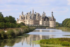 Château de Chambord reflected in the calm canal, surrounded by lush greenery on a bright day in the Loire Valley, France.
