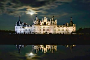 Château de Chambord illuminated at night under a glowing full moon, reflected perfectly in the still waters of the canal.