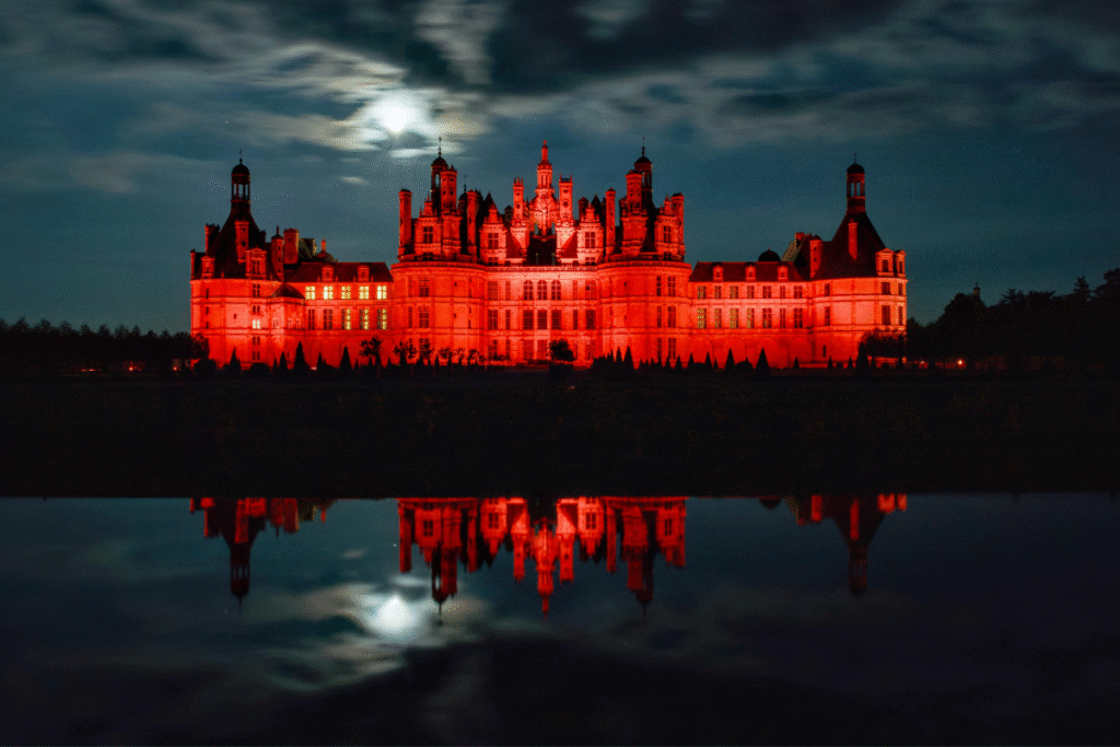 Château de Chambord iluminado em vermelho durante a noite, refletido na água sob a luz da lua cheia.