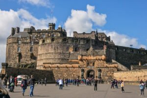Visitors exploring the Esplanade in front of the main entrance of Edinburgh Castle on a sunny day, with the iconic Half Moon Battery rising above.