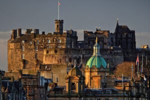 View of Edinburgh Castle rising behind the historic skyline at golden hour, with the green dome of the Bank of Scotland building in the foreground.