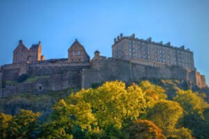 Edinburgh Castle seen from below, glowing in warm light above green and golden autumn trees against a clear sky.