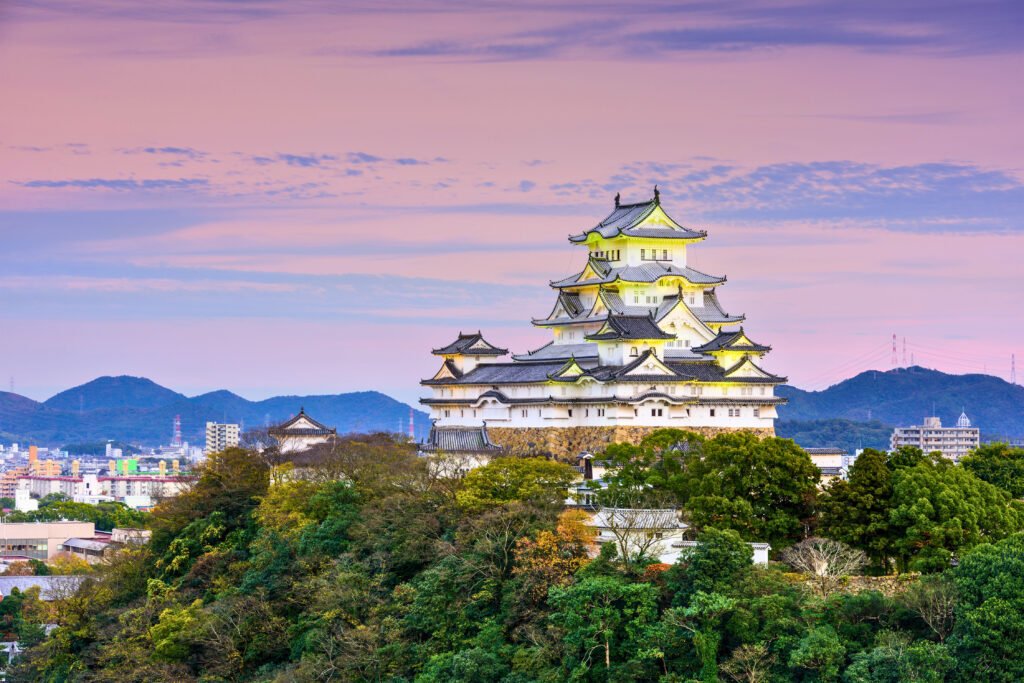 Himeji Castle in Japan illuminated by soft evening lights, standing majestically on a hilltop with a pink and purple sunset sky and distant mountains in the background.