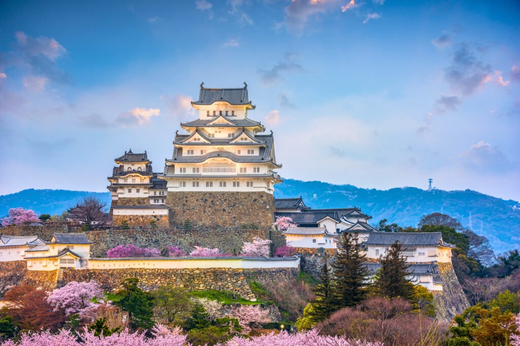 Majestic view of Himeji Castle at sunset with cherry blossoms in full bloom and distant mountains in the background under a soft blue and pink sky.