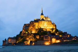 Mont-Saint-Michel glowing with evening lights against the blue twilight sky, iconic focus keyword displayed.