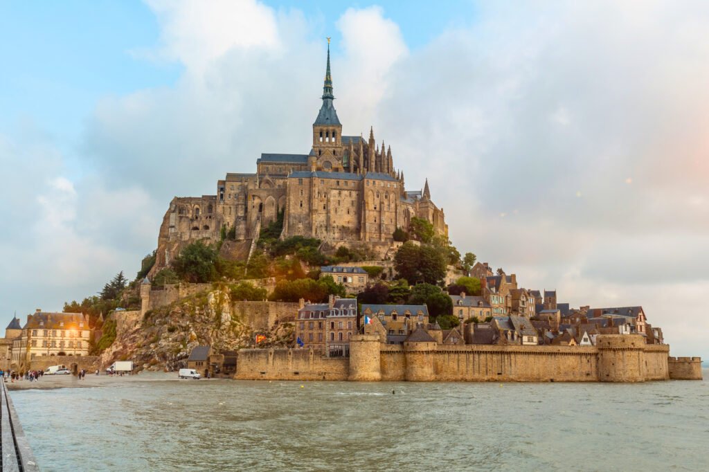 Mont Saint-Michel at high tide during sunset, with golden light illuminating the abbey and fortified village as seen from the causeway in Normandy, France.