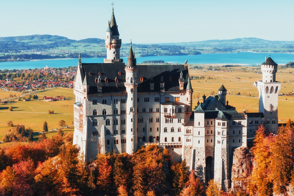 Uma vista de tirar o fôlego do Castelo de Neuschwanstein, cercado pela folhagem vibrante do outono, com campos ondulados e lagos azul-claros cintilando ao fundo na Baviera, Alemanha.