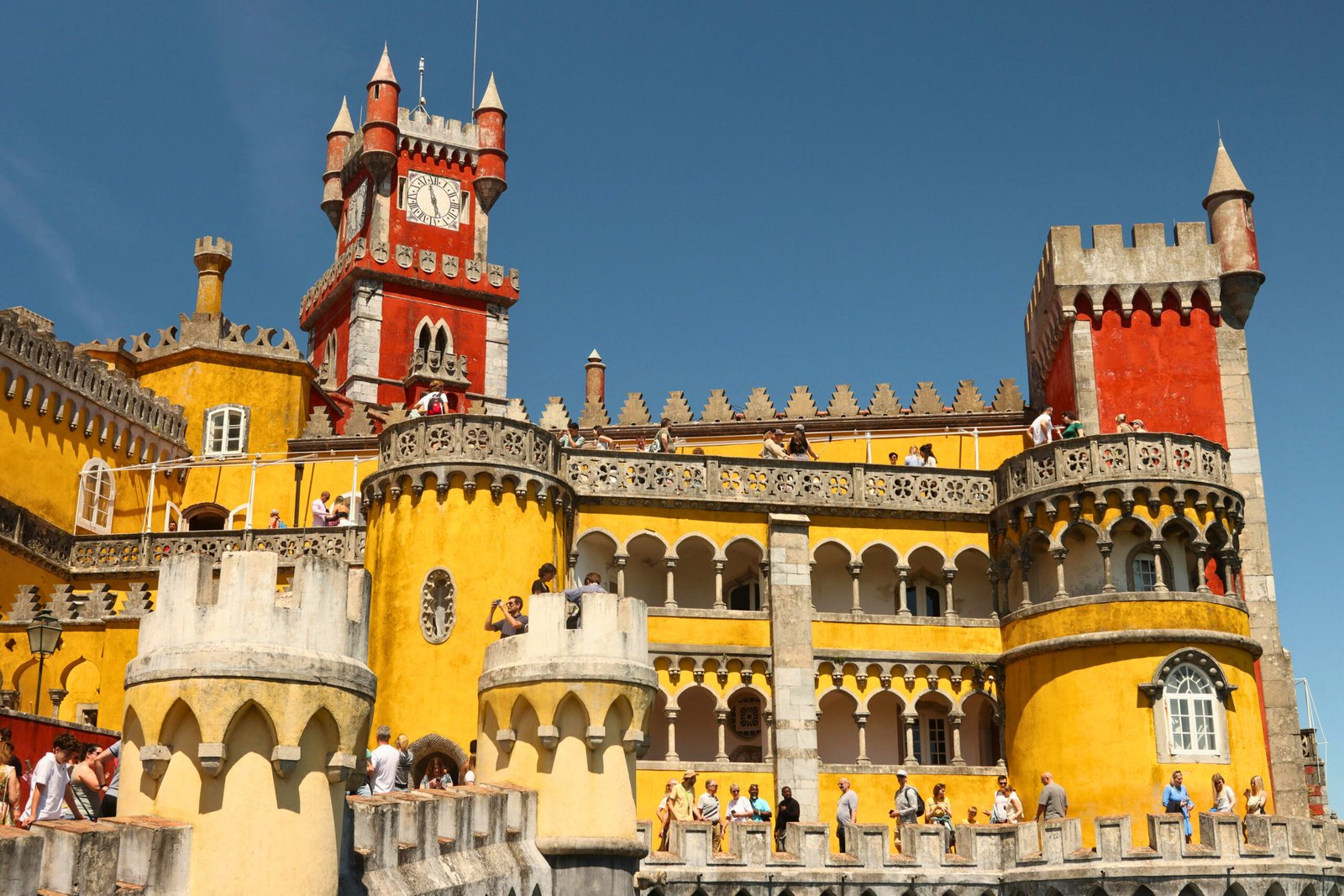 Tourists explore the vibrant façades and romantic towers of Pena Palace under clear summer skies.