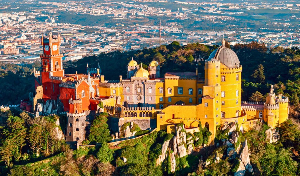 Aerial view of Pena Palace in Sintra, Portugal, showcasing its vibrant red and yellow Romanticist architecture atop a lush forested hill with the cityscape in the background.