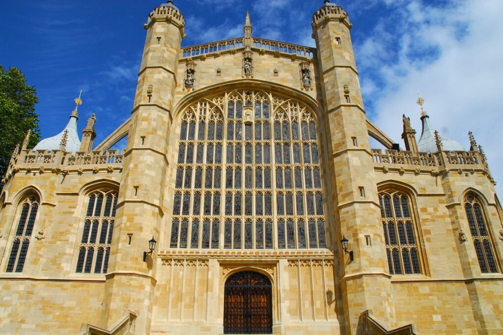 La impresionante fachada de la Capilla de San Jorge en el Castillo de Windsor, que muestra su intrincada arquitectura gótica perpendicular, grandes vitrales de colores y detallados adornos en piedra, todo bajo un cielo azul brillante.
