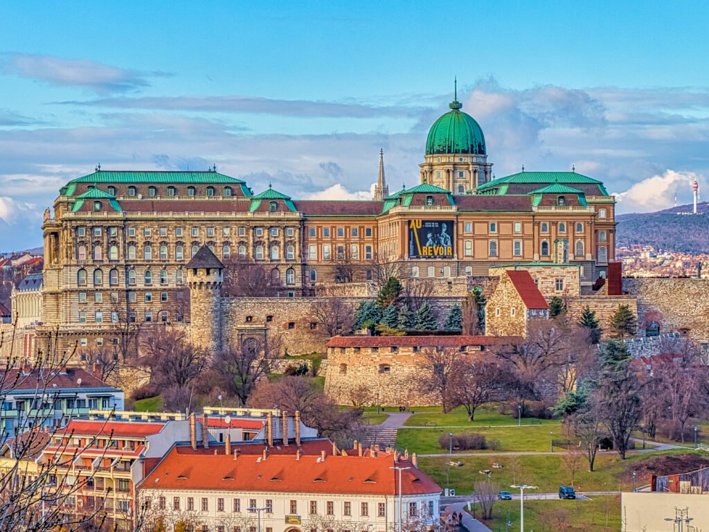 Colorful daytime view of Buda Castle in Budapest, Hungary, featuring medieval fortifications, Baroque architecture, and the iconic green dome against a blue sky.
