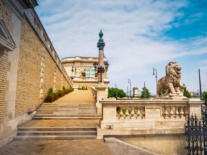 Buda Castle lion statue guarding historic Budapest staircase, blue sky above.
