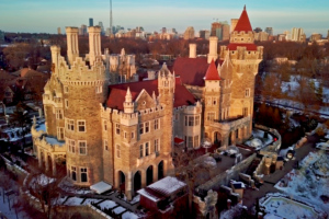 Aerial view of Casa Loma in winter with snow-covered rooftops and garden, set against the Toronto skyline at sunset.