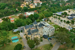 High-angle aerial view of Castelo de Itaipava, showing the full castle complex, outdoor pool, gardens, parking area, and surrounding residential neighborhood.