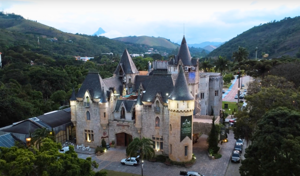 Aerial view of Castelo de Itaipava in Brazil, a Neo-Gothic castle surrounded by lush green hills and palm trees, lit up at dusk.