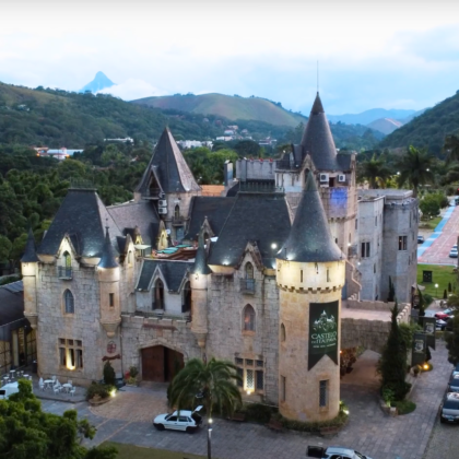 Aerial view of Castelo de Itaipava in Brazil, a Neo-Gothic castle surrounded by lush green hills and palm trees, lit up at dusk.