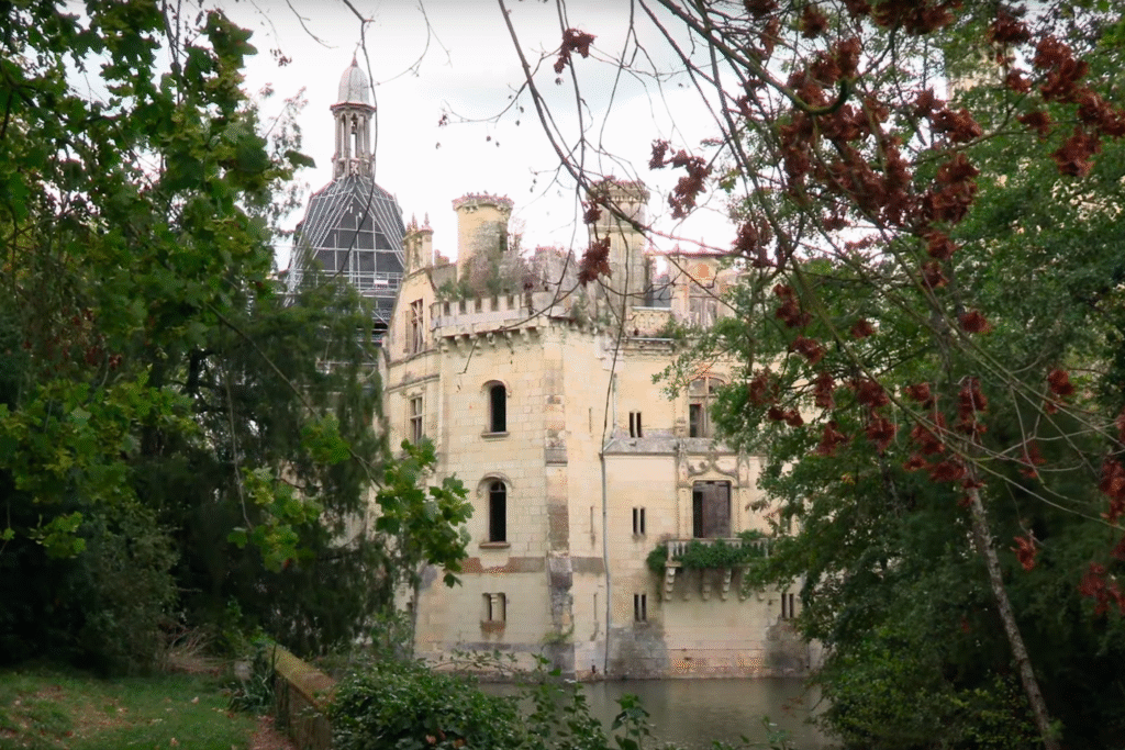 Partial view of la Mothe-Chandeniers through autumn foliage, highlighting its turreted walls, overgrown rooftop, and restoration scaffolding on the central dome.
