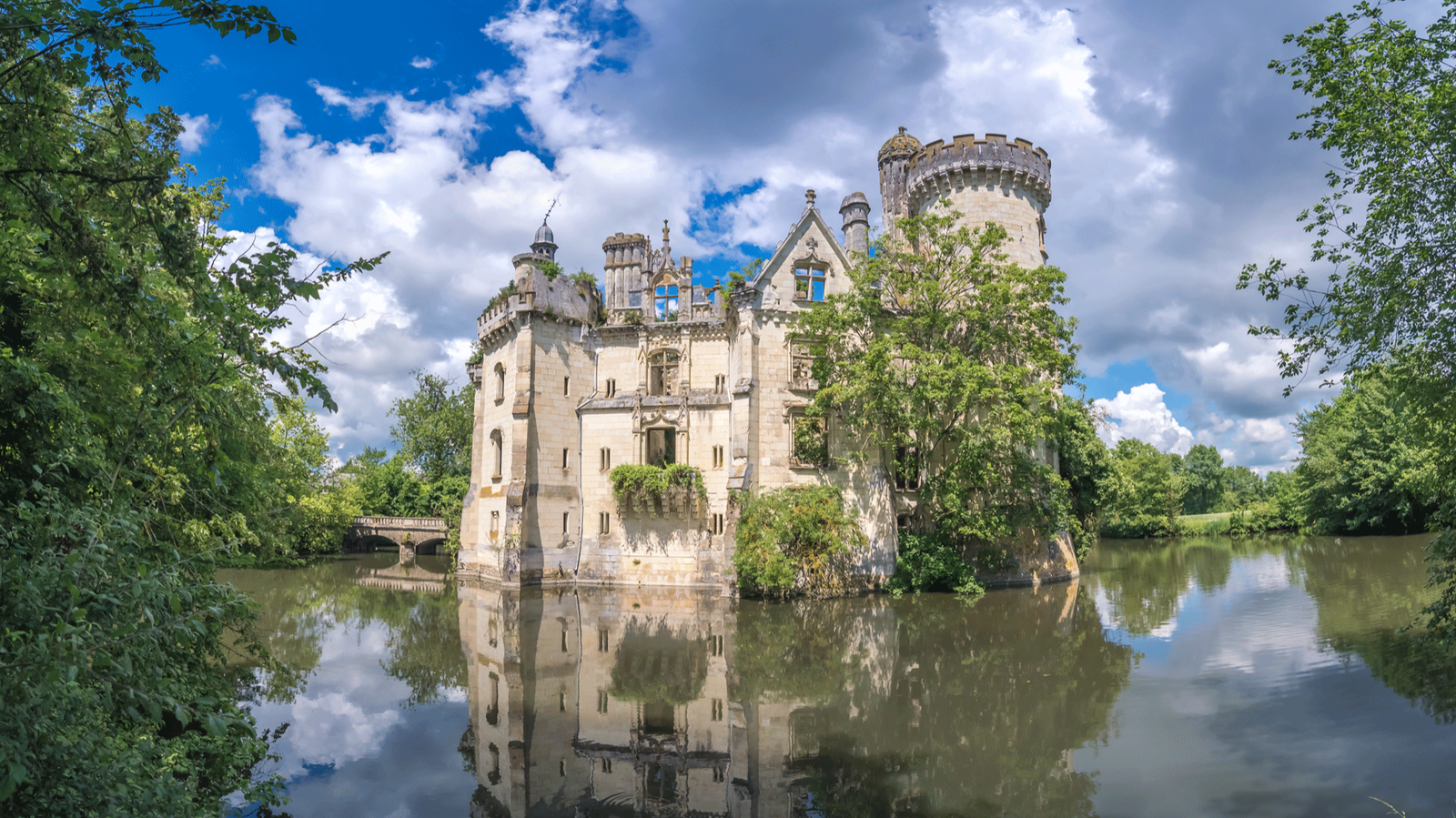 La Mothe-Chandeniers reflecting in a calm moat under a bright summer sky, surrounded by vibrant greenery and framed by forested banks.