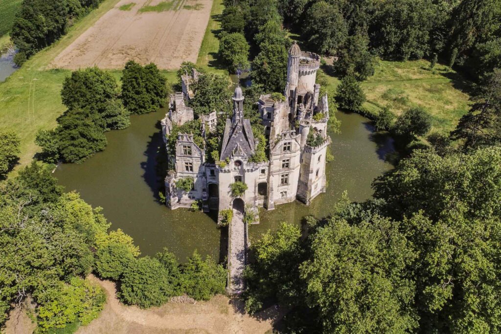 Aerial view of the Castle of la Mothe-Chandeniers fully surrounded by a moat, with its Neo-Gothic ruins overtaken by greenery and forest on all sides.