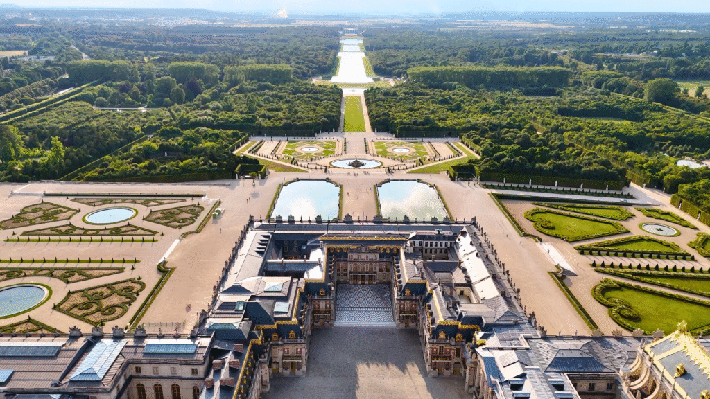 Aerial view of the Palace of Versailles and its symmetrical gardens stretching toward the Grand Canal, showcasing the scale and design of the Baroque masterpiece.