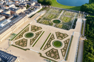 Aerial view of the meticulously designed parterre gardens of the Palace of Versailles, featuring symmetrical patterns, circular fountains, and tree-lined walkways near the city.