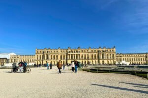 Panoramic view of the Palace of Versailles on a sunny day, with visitors walking across the gravel forecourt and the grand Baroque façade bathed in golden light.