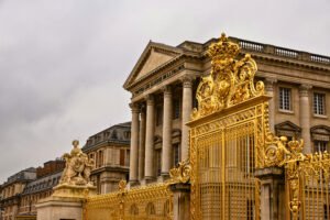 Golden gate of the Palace of Versailles in front of the classical colonnaded building inscribed with “À toutes les gloires de la France,” under a cloudy sky.