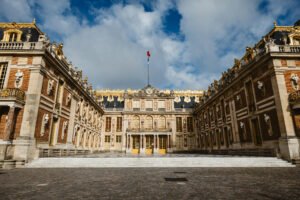 The Marble Courtyard of the Palace of Versailles, featuring ornate red-brick and stone façades, gilded details, and classical sculptures beneath a dramatic sky.