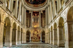 Interior of the Royal Chapel at the Palace of Versailles, featuring fluted Corinthian columns, ornate white stone carvings, and a gilded altar and pipe organ.