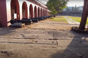 Row of historical cannons displayed along the red-arched museum corridor at Fort Jesus, Mombasa, surrounded by a spacious courtyard and shaded trees.