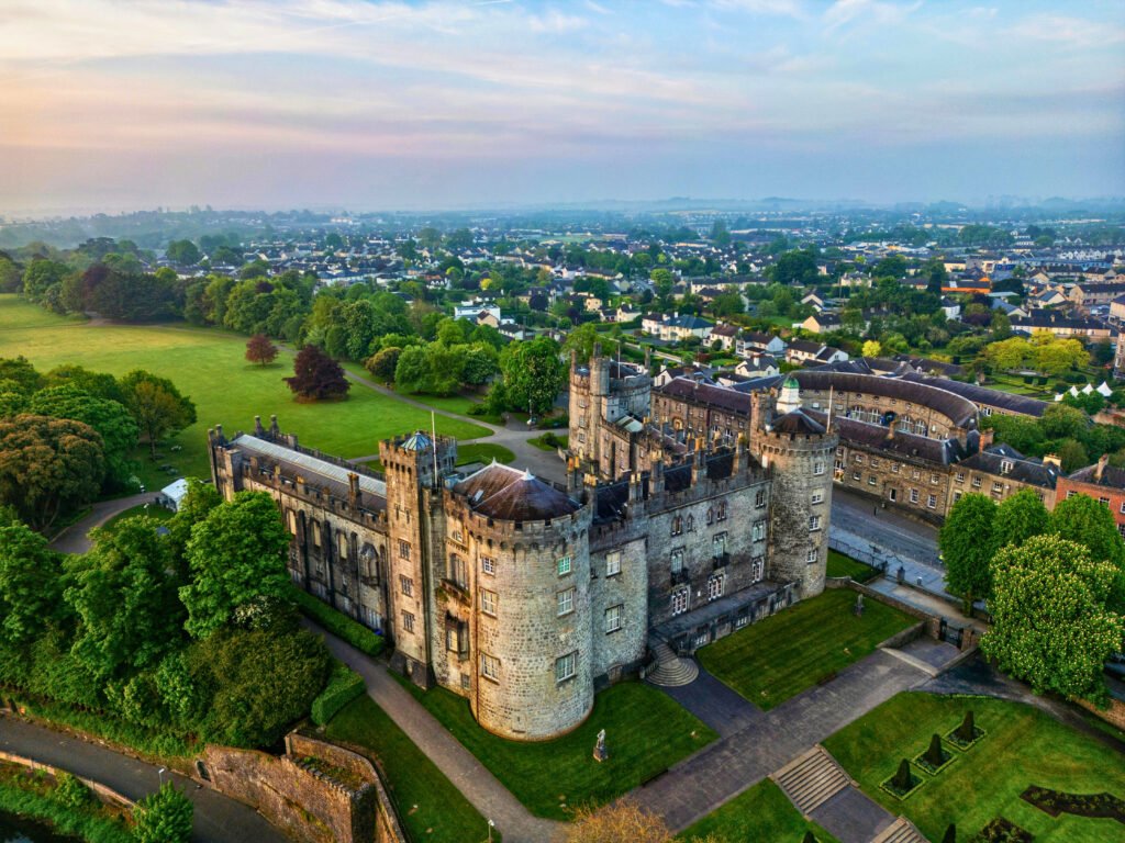 Aerial view of Kilkenny Castle in Ireland, showcasing its medieval architecture, manicured gardens, and surrounding cityscape at sunset.