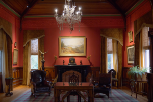 Elegant Victorian-style room inside Larnach Castle featuring crimson walls, antique furniture, a grand chandelier, and a central fireplace with period décor.
