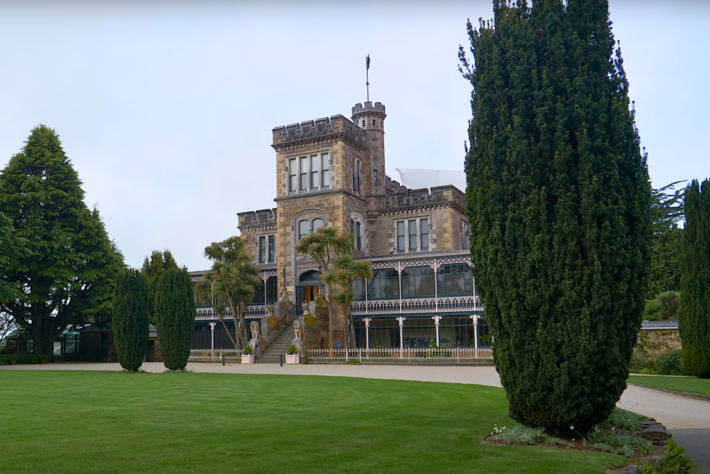 Wide view of Larnach Castle’s front façade with its central tower, Victorian glass veranda, and lush green lawn bordered by manicured trees in Dunedin, New Zealand.