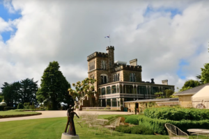 Side view of Larnach Castle with outdoor seating and gardens in the foreground, showcasing the castle’s veranda, tower, and landscaped grounds under a partly cloudy sky.