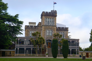 Frontal view of Larnach Castle with its central tower, New Zealand flag flying above, and detailed Victorian-era architecture surrounded by manicured gardens.