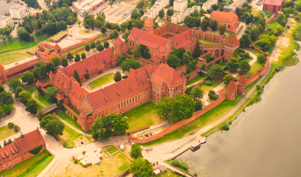 Aerial view of Malbork Castle in Poland, showcasing its massive red-brick Gothic architecture, defensive walls, and riverside location along the Nogat River.