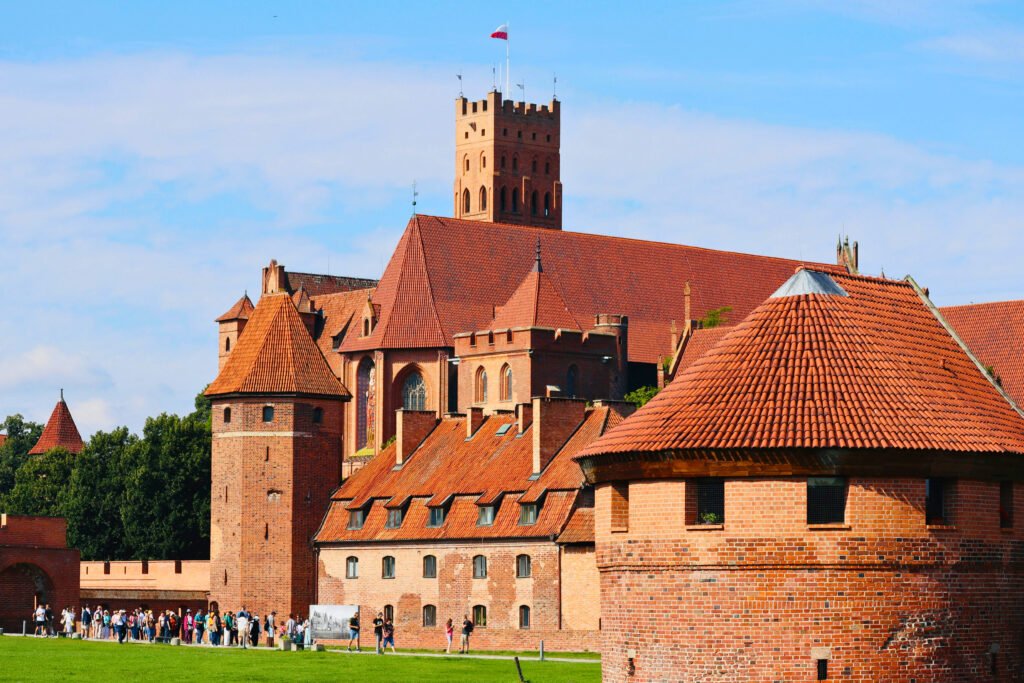 Visiteurs marchant près des tours fortifiées et des toits en tuiles rouges de la structure gothique en brique du château de Malbork sous un ciel bleu éclatant.