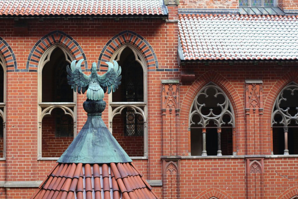 Gros plan sur des fenêtres à arches gothiques et une sculpture d'aigle en cuivre au sommet d'un toit en tuiles dans la cour intérieure du château de Malbork, mettant en valeur ses détails gothiques en brique.