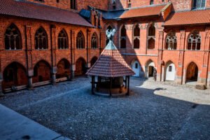 The inner courtyard of Malbork Castle featuring a central well with a tiled canopy and Gothic arched walkways surrounding the cobblestone square.
