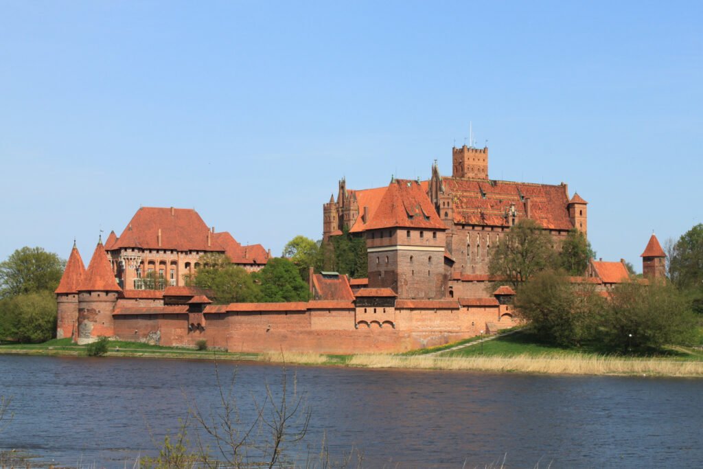 Wide-angle view of Malbork Castle’s towering red-brick structures and defensive walls overlooking the calm waters of the Nogat River under a clear blue sky.