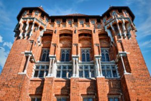 Close-up upward view of Malbork Castle’s richly detailed red-brick facade featuring arched Gothic windows, stone columns, and decorative corbels under a blue sky.
