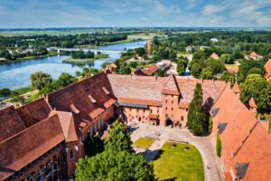 Aerial view of Malbork Castle’s inner courtyard and red-tiled rooftops, with the Nogat River, lush greenery, and rural Polish countryside in the distance.