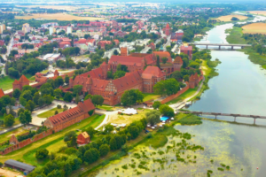 Wide aerial view of Malbork Castle in Poland, with its sprawling red-brick complex set against the Nogat River and the town of Malbork in the background.