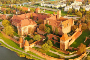 Aerial view of Malbork Castle in autumn, highlighting the red-brick Gothic complex, defensive towers, and surrounding greenery near the Nogat River.
