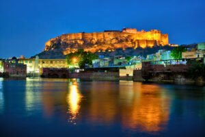 Mehrangarh Fort illuminated at night, reflecting in the lake below with the surrounding cityscape of Jodhpur under a deep blue sky.
