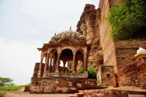 Stone cenotaph pavilion near the base of Mehrangarh Fort in Jodhpur, India, framed by towering fort walls and lush greenery.